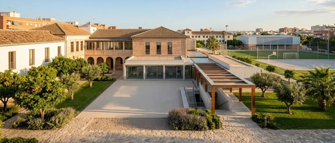 Fotografía realista de la arquitectura exterior de un centro educativo en Valencia, mostrando el patio central, zonas verdes y campos deportivos bajo una luz de atardecer.
